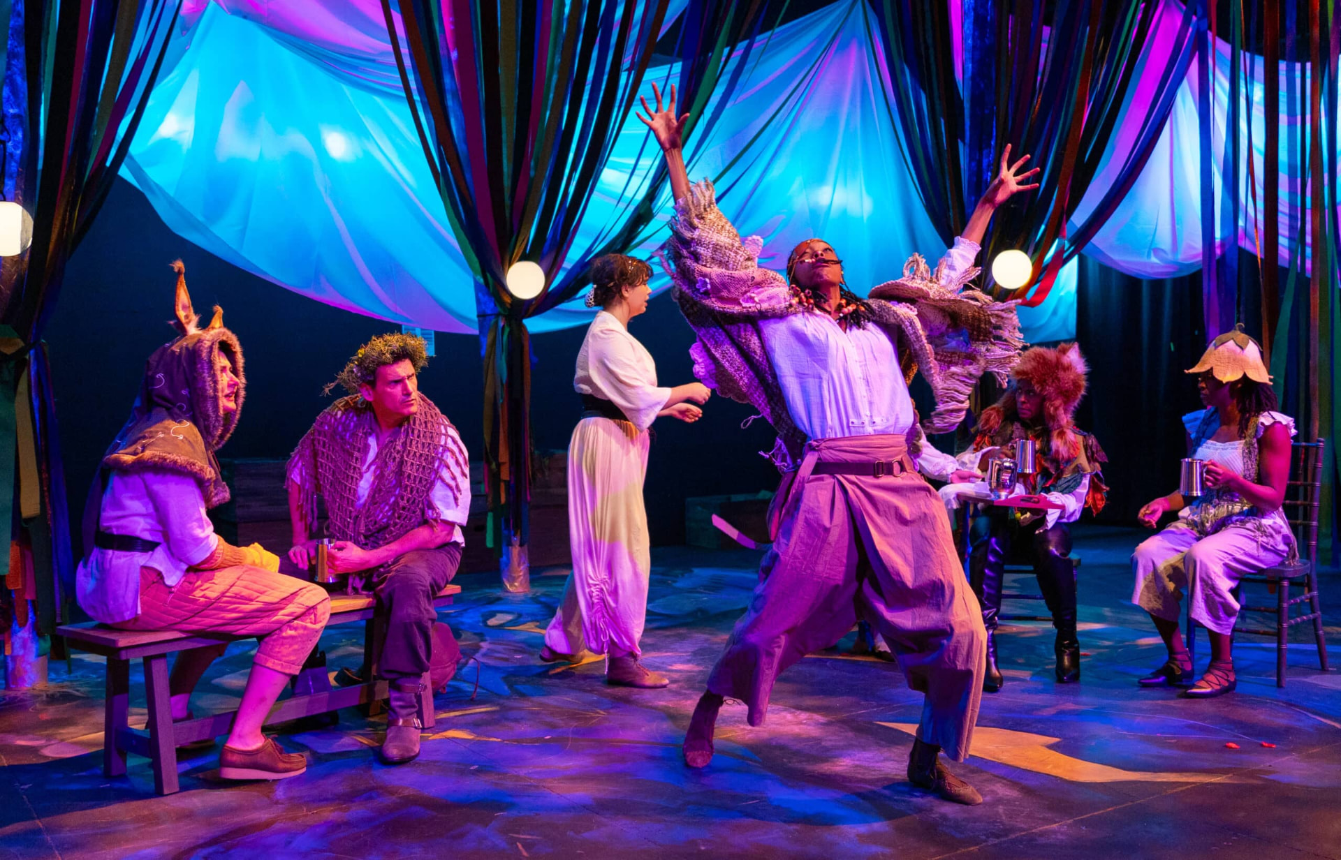 The ensemble of American Stage's production of The Figs performs in vibrant costumes under blue and purple stage lighting.