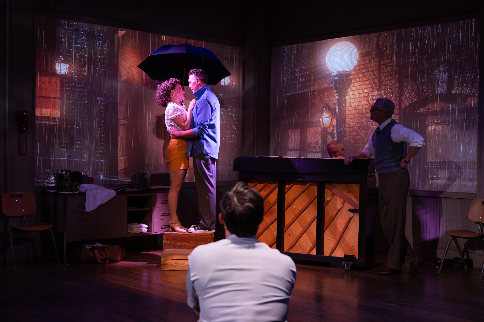 Two actors stand under an umbrella in front of projected rain-soaked street scenes