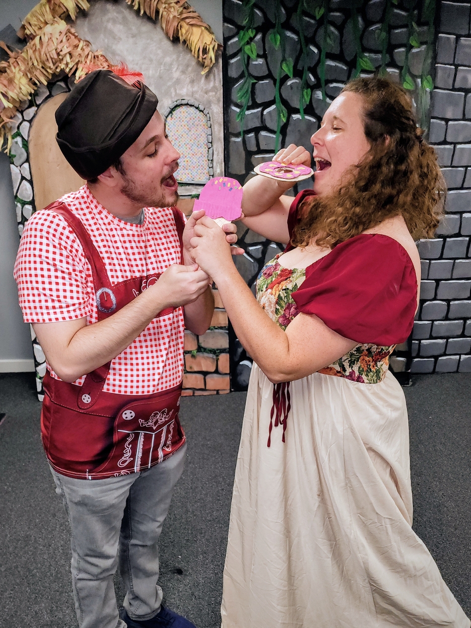 Two actors mock-fight over paper pastries in a bakery scene.