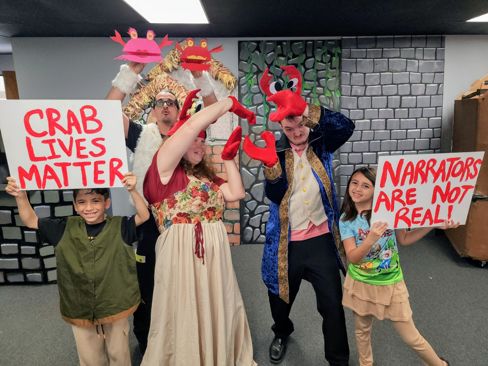 Cast members hold 'Crab Lives Matter' and 'Narrators Are Not Real' signs in costume.