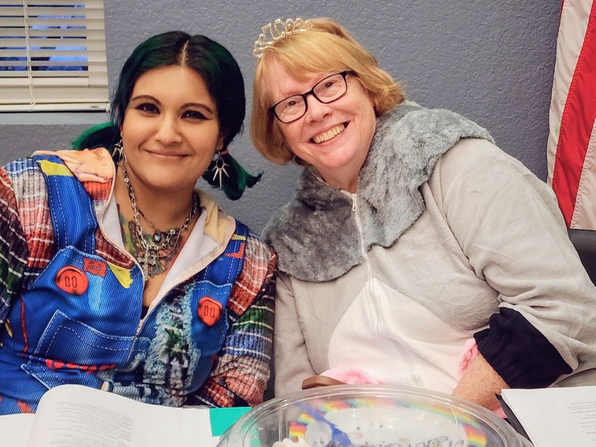 Two Beyond the Wings Theater cast members smile side by side in costume, one in a patchwork doll outfit and the other in a fuzzy costume and tiara, with scripts on the table.