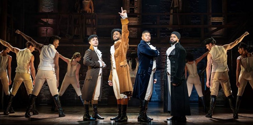 The principal cast of the Hamilton North American tour stands in a line at center stage, the performer in the tan coat pointing upward, ensemble members posed with arms outstretched around them.